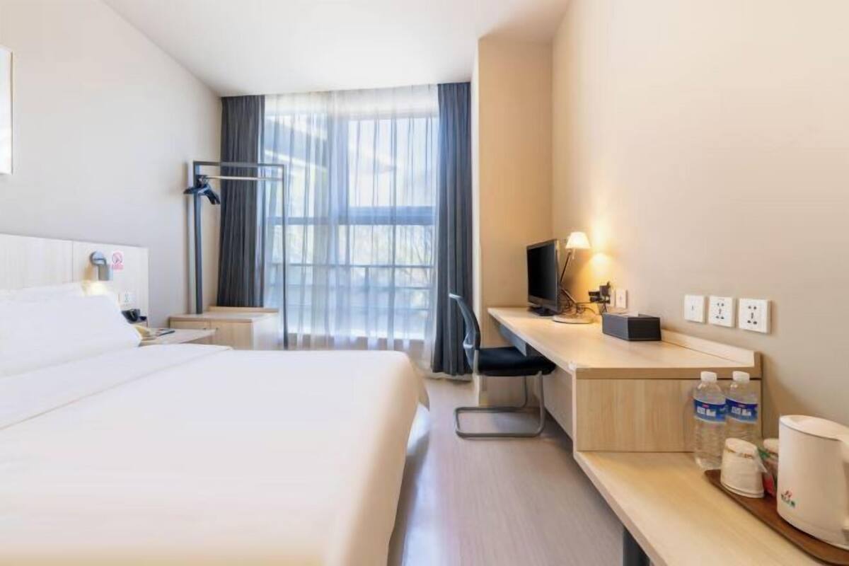 A hotel room with a bed, a desk with a computer, a chair, a TV, and a window with curtains.