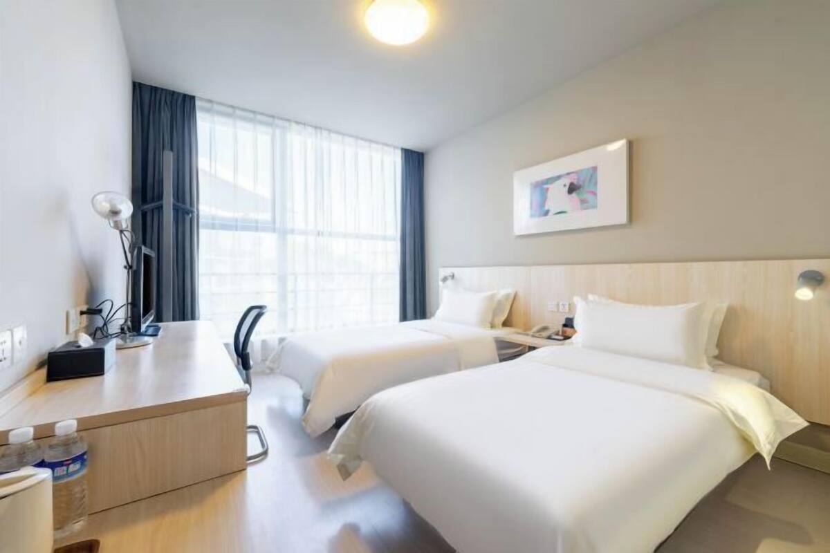 A hotel room with two beds, a desk with a lamp, a chair, and a window with curtains.