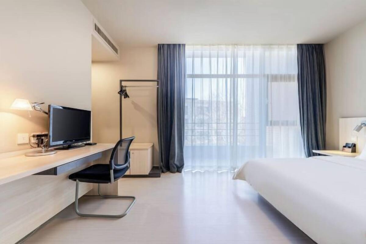 A hotel room with a bed, a desk with a computer, a chair, a window with curtains, and a view of the city.