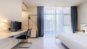 Quarto de hotel com uma cama, uma escrivaninha com computador, uma cadeira, uma janela com cortinas e vista para a cidade.