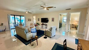 A spacious living room with a sofa, armchairs, a coffee table, and a TV. There are ceiling fans, a balcony with a view, and a hallway leading to other rooms. - Beachcomber Lodge in Fort Myers Beach (Fort Myers Beach)