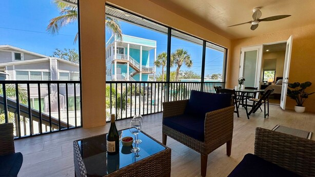 A balcony with wicker furniture, a table with a bottle and glasses, and a view of a blue building and palm trees. - Beachcomber Lodge in Fort Myers Beach (Fort Myers Beach)
