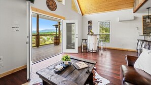 A spacious living area with a wooden ceiling, a large window overlooking a scenic landscape, a rustic coffee table, and a leather sofa. - Tennessee Whiskey Cabin- Lake and Mtn Views, Covered Deck, Game Room, Pool Table, BBQ, Pets (Sevierville)