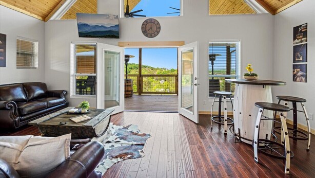 A spacious living room with a leather couch, a coffee table, and a rug. There is a wooden deck leading to a balcony with a view of the mountains. - Tennessee Whiskey Cabin- Lake and Mtn Views, Covered Deck, Game Room, Pool Table, BBQ, Pets (Sevierville)