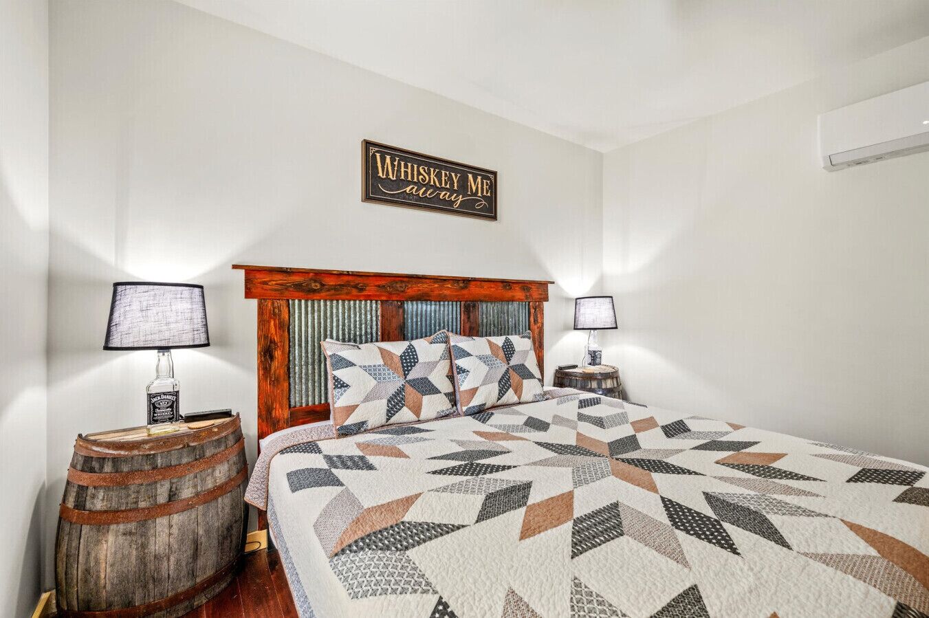Une chambre avec une tête de lit en bois, une couvre-lit matelassé, deux lampes de chevet, un tonneau à whisky et une pancarte « Whiskey Me » accrochée au mur.