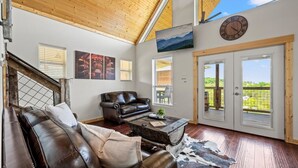 A spacious living room with a brown leather sofa, a wooden coffee table, and a large window offering a view of the outdoors. - Tennessee Whiskey Cabin- Lake and Mtn Views, Covered Deck, Game Room, Pool Table, BBQ, Pets (Sevierville)