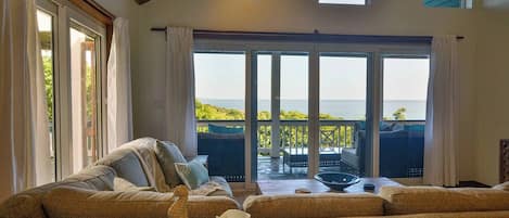 A cozy living room with a large sofa, a coffee table, and a view of the sea through large sliding glass doors.