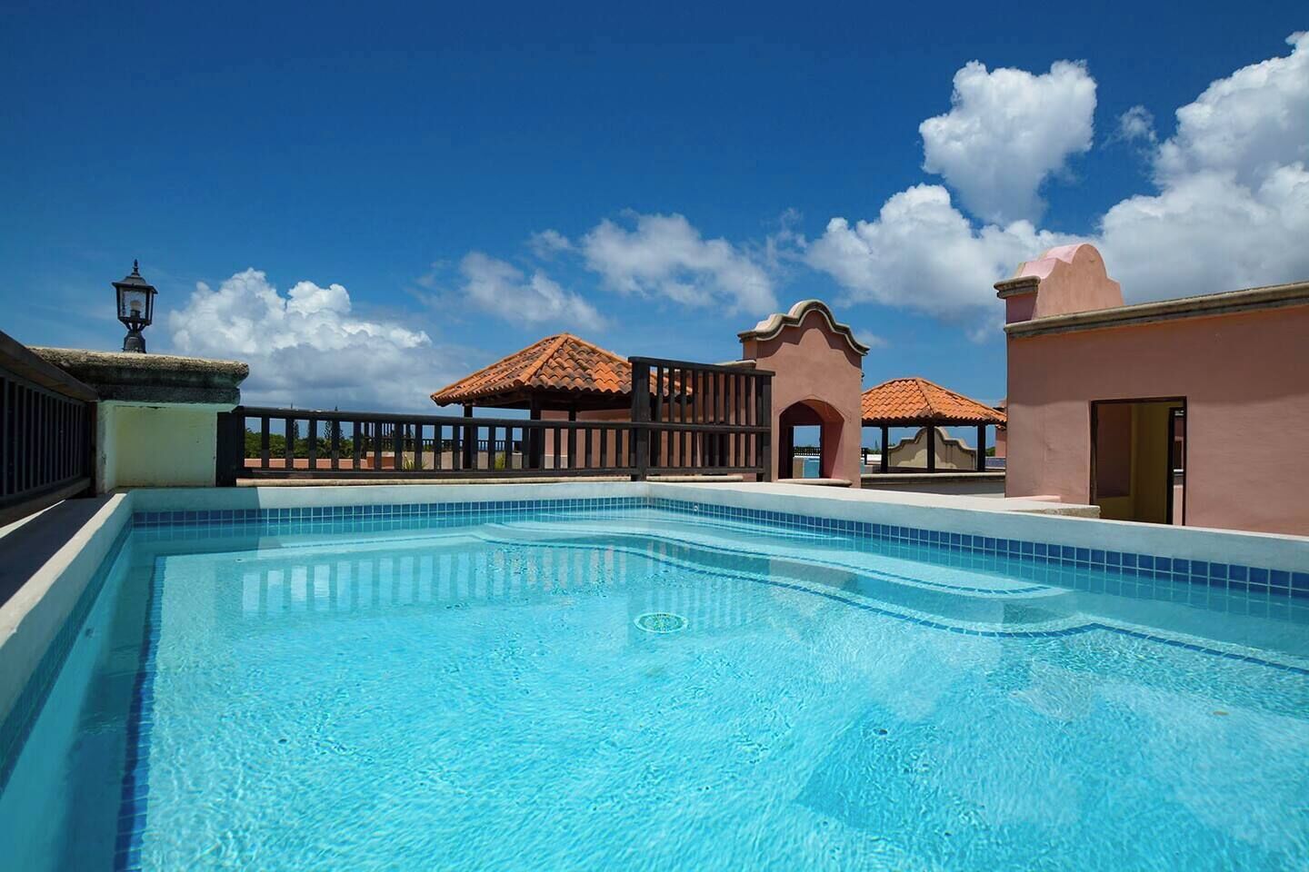 A swimming pool with a tiled deck, a gazebo, and a building with a pink facade.