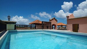 A swimming pool with a tiled deck, a gazebo, and a building with a pink facade. - Mission Gate 1A,  1-bedroom condo by Island House Vacation Rentals (West Bay)