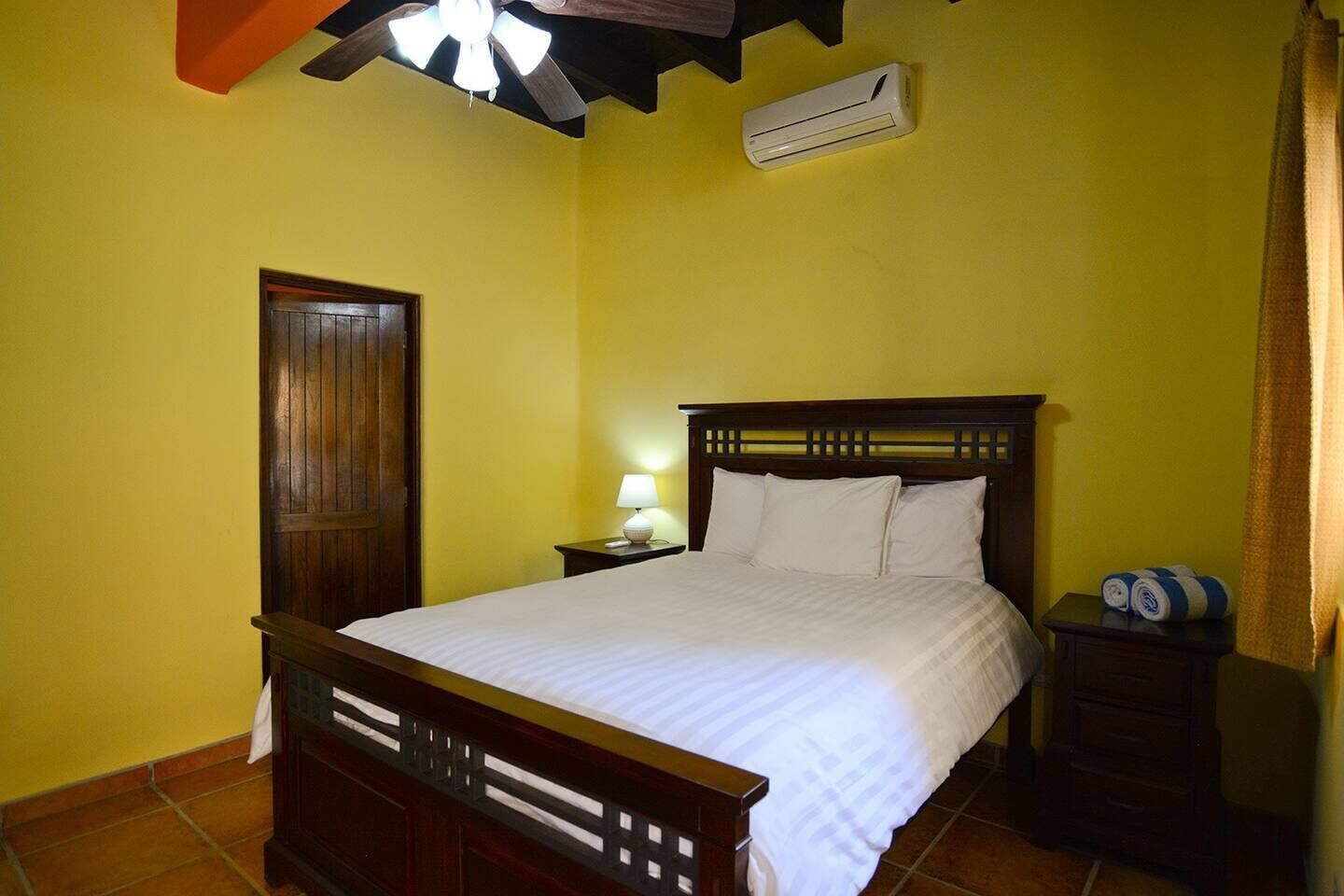 A bedroom with a wooden bed, a nightstand with a lamp, an air conditioner, and a door.