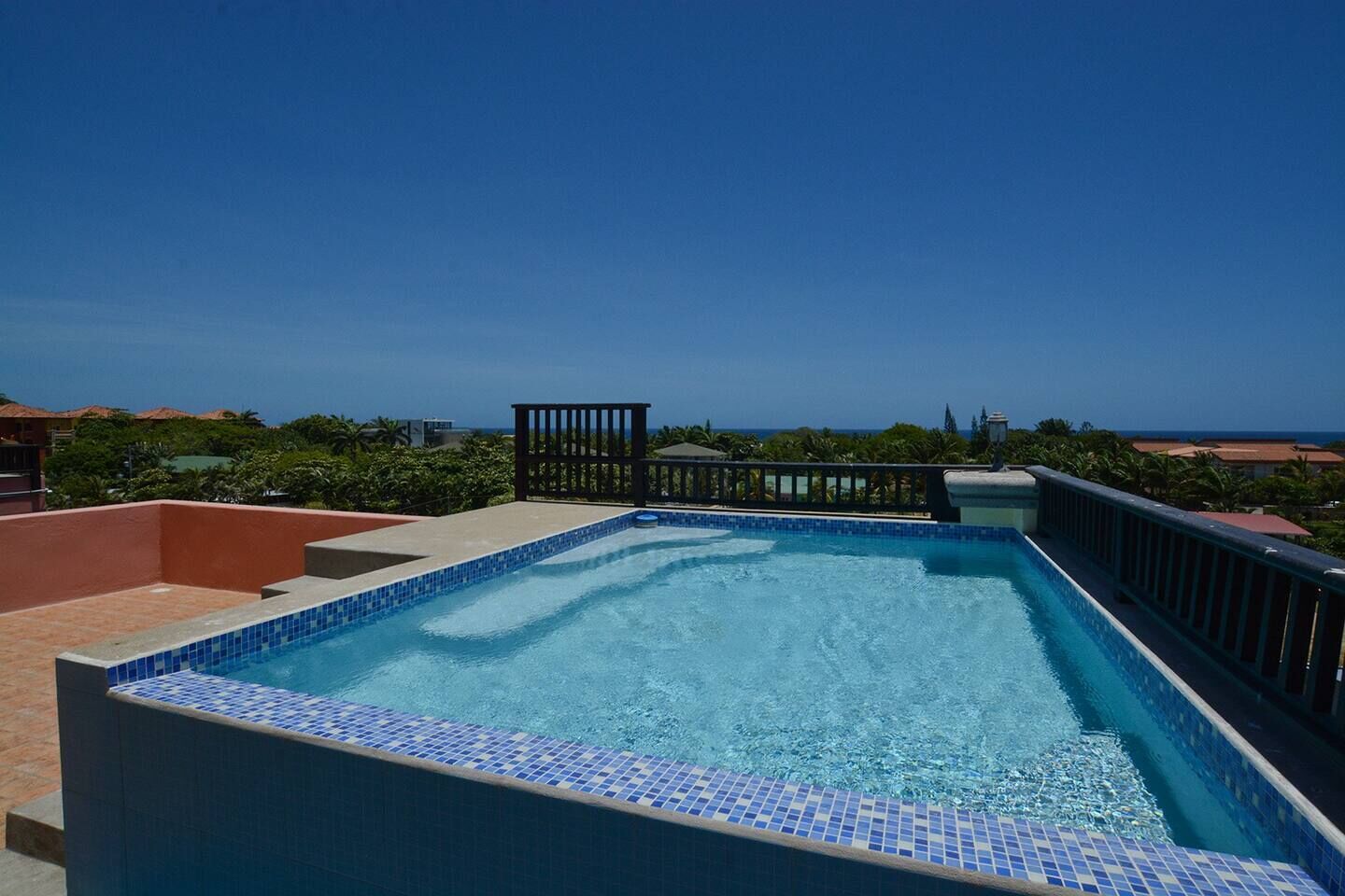 Una piscina rectangular con mosaicos azules, rodeada por un muro de ladrillo bajo y una barandilla negra.