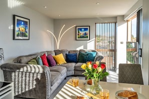 Un salon moderne avec un canapé gris, des coussins colorés, une table à manger agrémentée d’un vase de fleurs et deux œuvres d’art encadrées accrochées au mur.