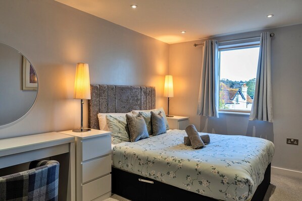 A bedroom with a bed, bedside lamps, a dresser, a mirror, and a window with curtains. - Apartment in Teignmouth With Sea Views (Teignmouth)