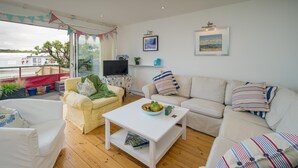 Un salon chaleureux avec un canapé blanc, un fauteuil jaune et une table basse blanche. On y trouve un téléviseur posé sur un meuble, une fenêtre avec des rideaux et une vue sur l’extérieur.