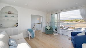 Un vaste espace de vie doté d’un plancher en bois, d’un canapé blanc, d’un canapé bleu et d’un fauteuil vert. Un balcon offre une vue sur la plage et les bateaux.