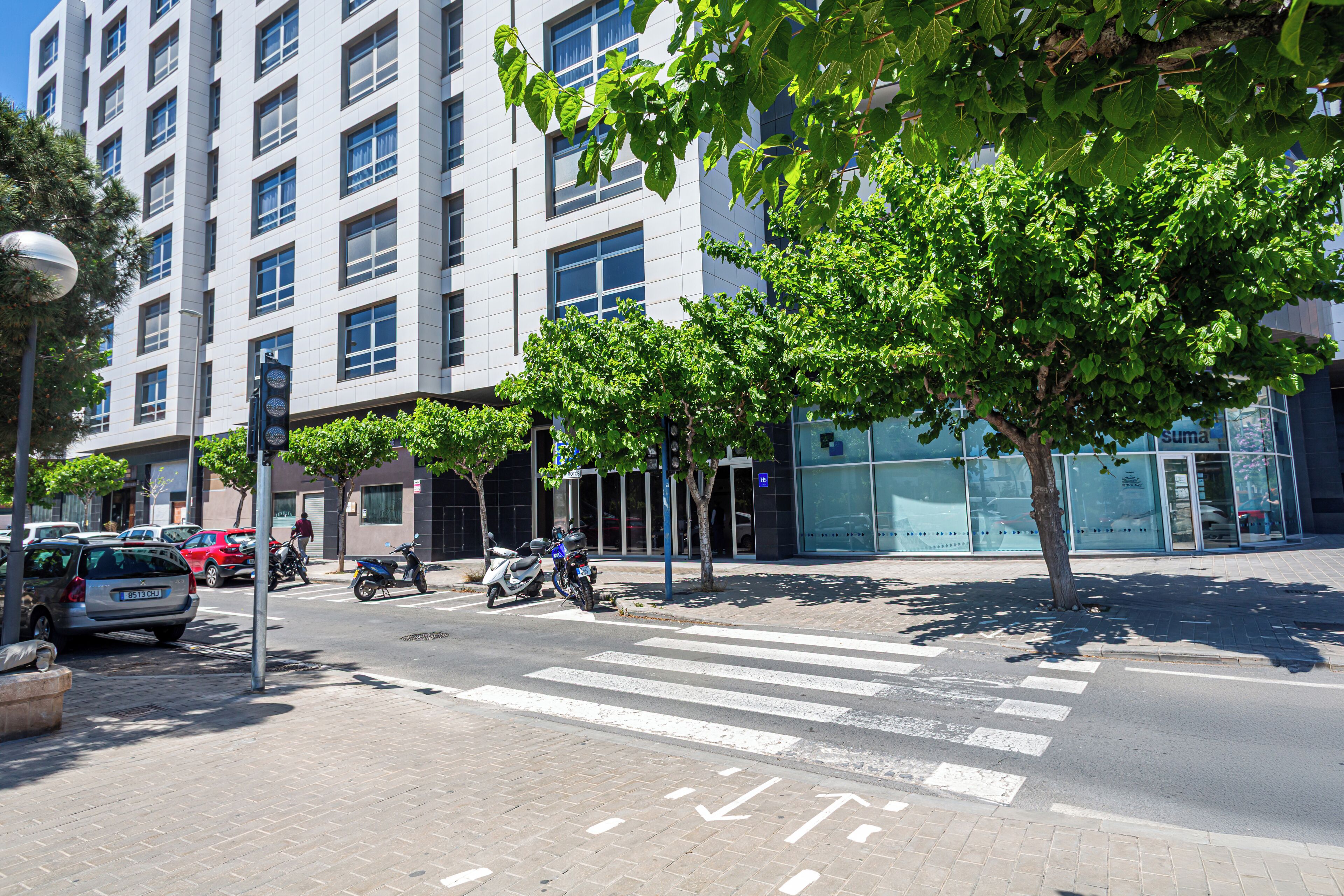 Un edificio moderno con un gran muro de cristal, un paso de peatones y vehículos estacionados.