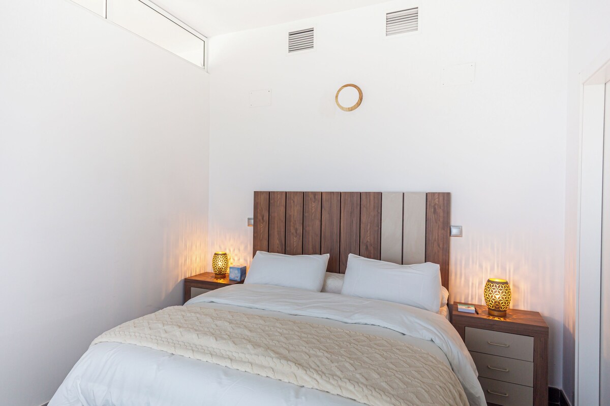 Una cama bien hecha con cabecera de madera, dos lámparas de noche y un pequeño espejo redondo en la pared.