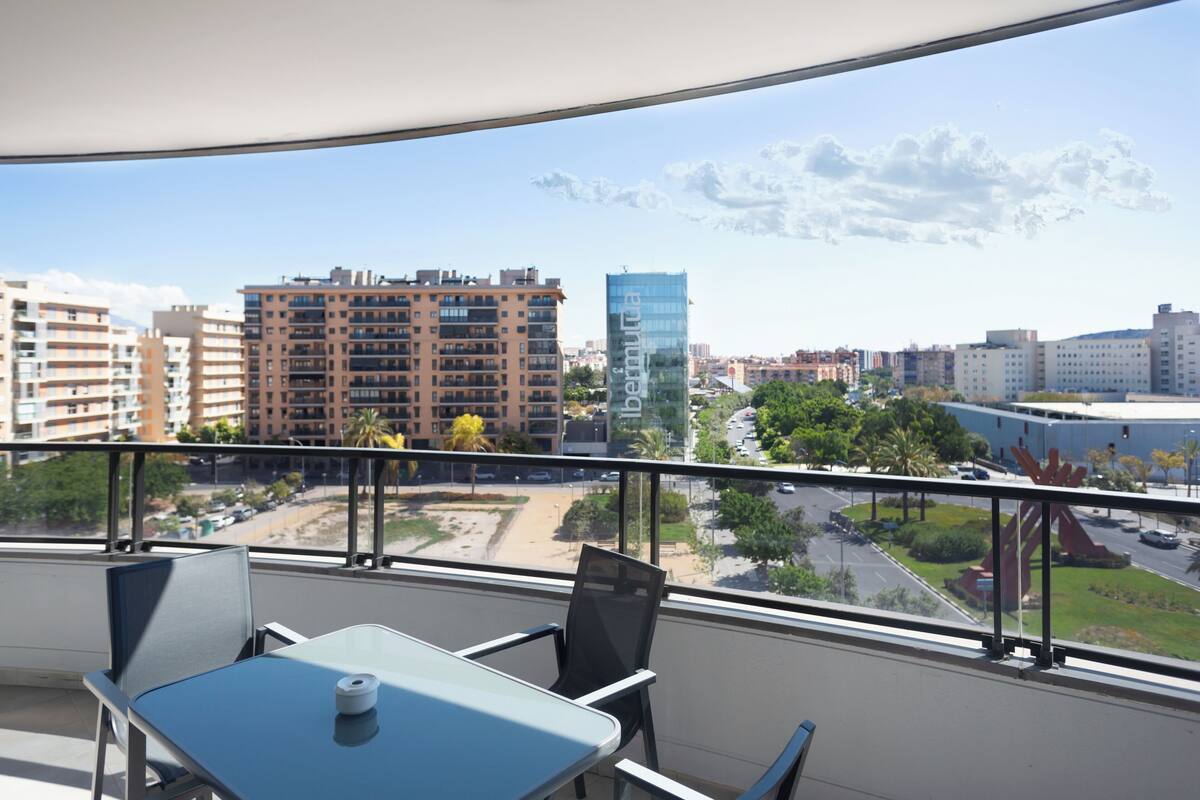 A balcony with a table and chairs, overlooking a cityscape with buildings and a road.