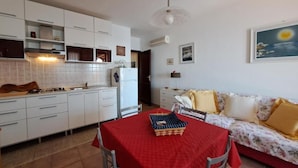 A compact living space with a kitchenette, a dining area with a red tablecloth, and a sofa with floral upholstery. - Casa Marina di Castelsardo (Castelsardo)