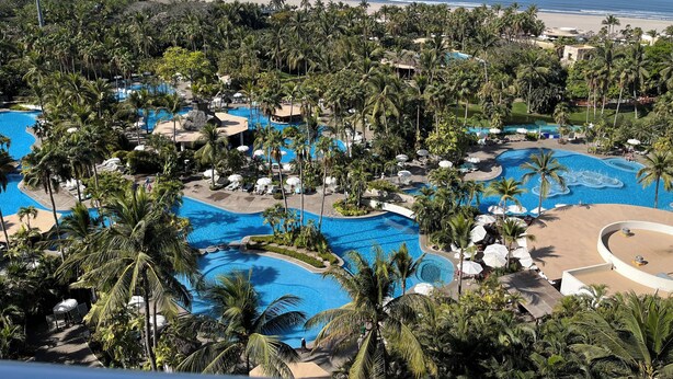 Ein Resort mit mehreren Schwimmbecken, umgeben von Palmen und Blick auf einen Strand.