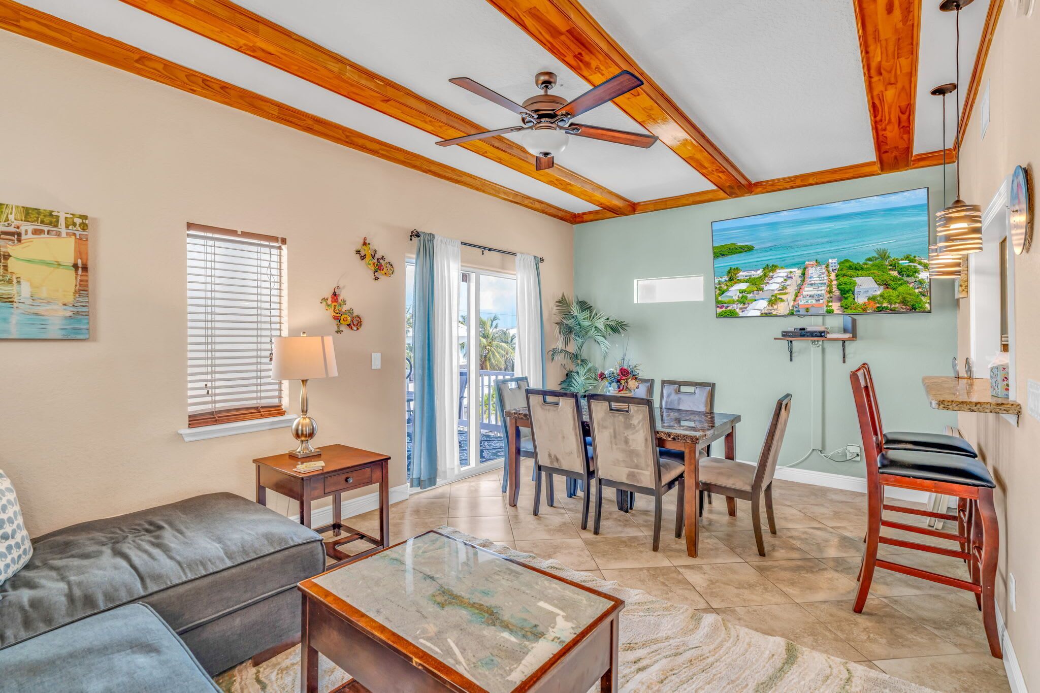 A spacious living room with a ceiling fan, a dining area with chairs and a table, and a large window with blinds.