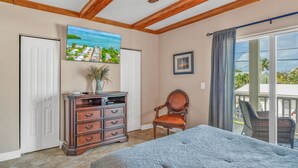 A bedroom with a bed, a dresser, a chair, a painting, and a balcony with a view.