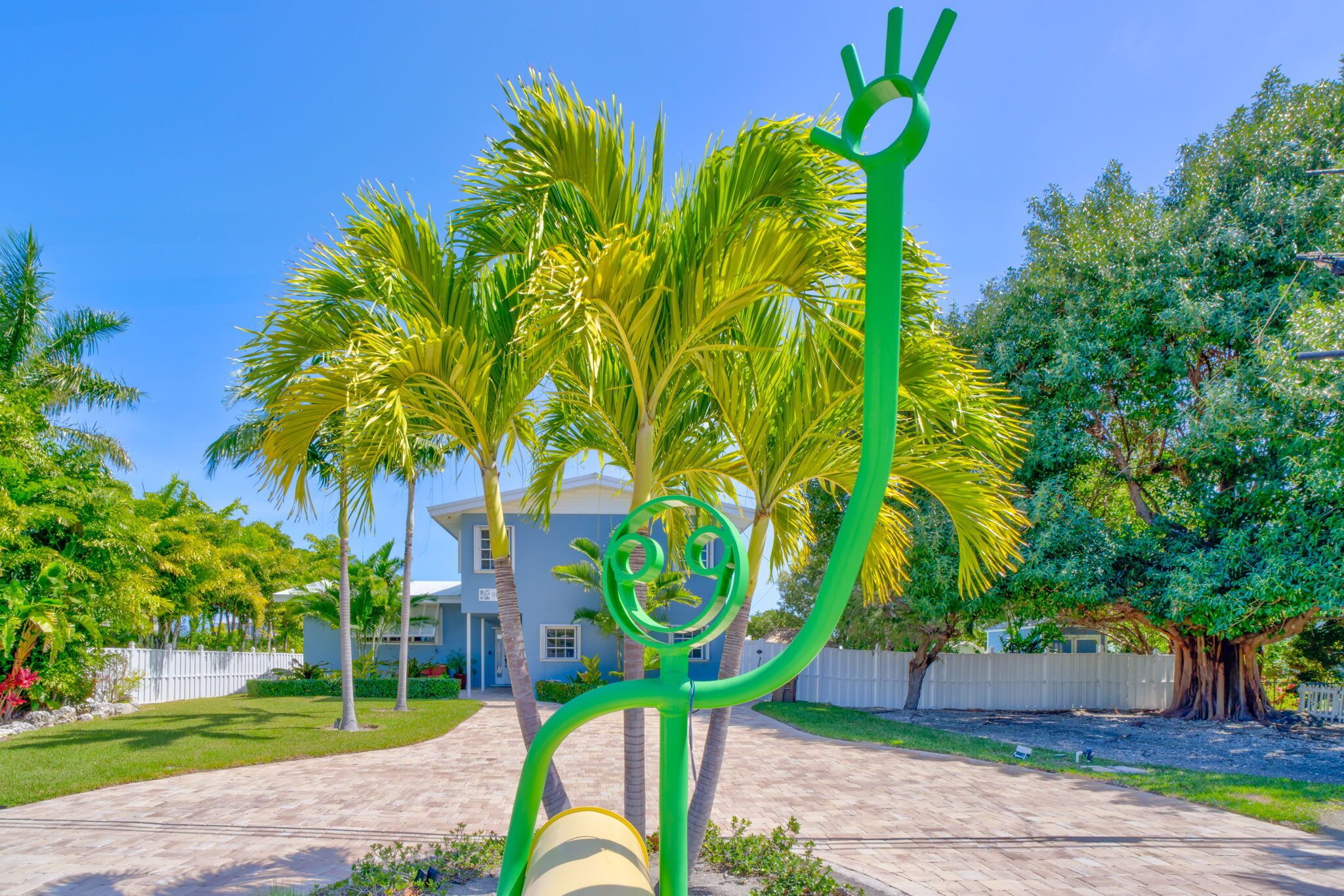 Zona residencial con camino de entrada pavimentado, palmeras y una casa con cerca blanca.