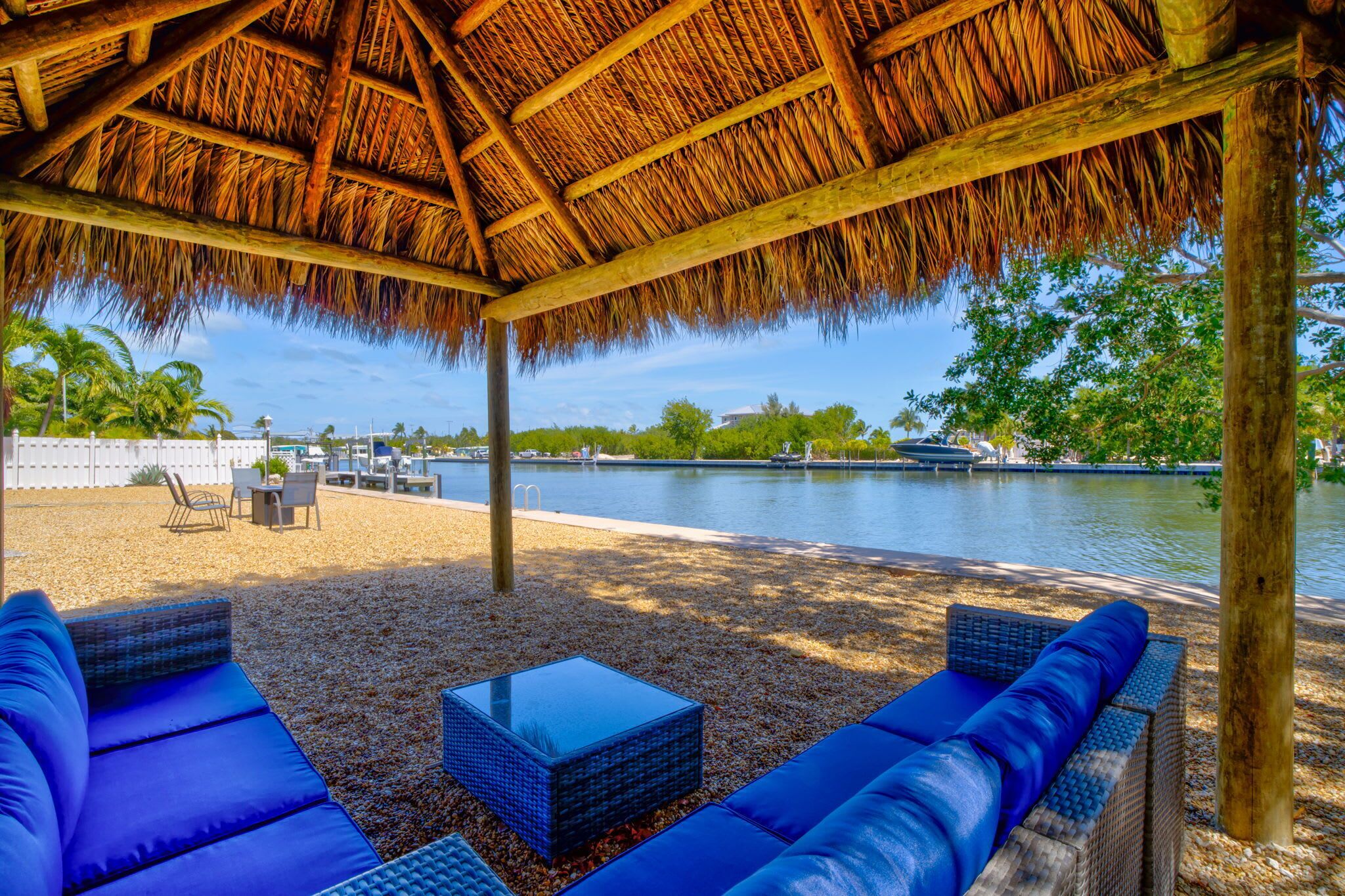 A covered outdoor seating area with blue cushions and a table, overlooking a body of water and greenery.