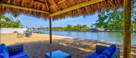 A covered outdoor seating area with blue cushions and a table, overlooking a body of water and greenery.
