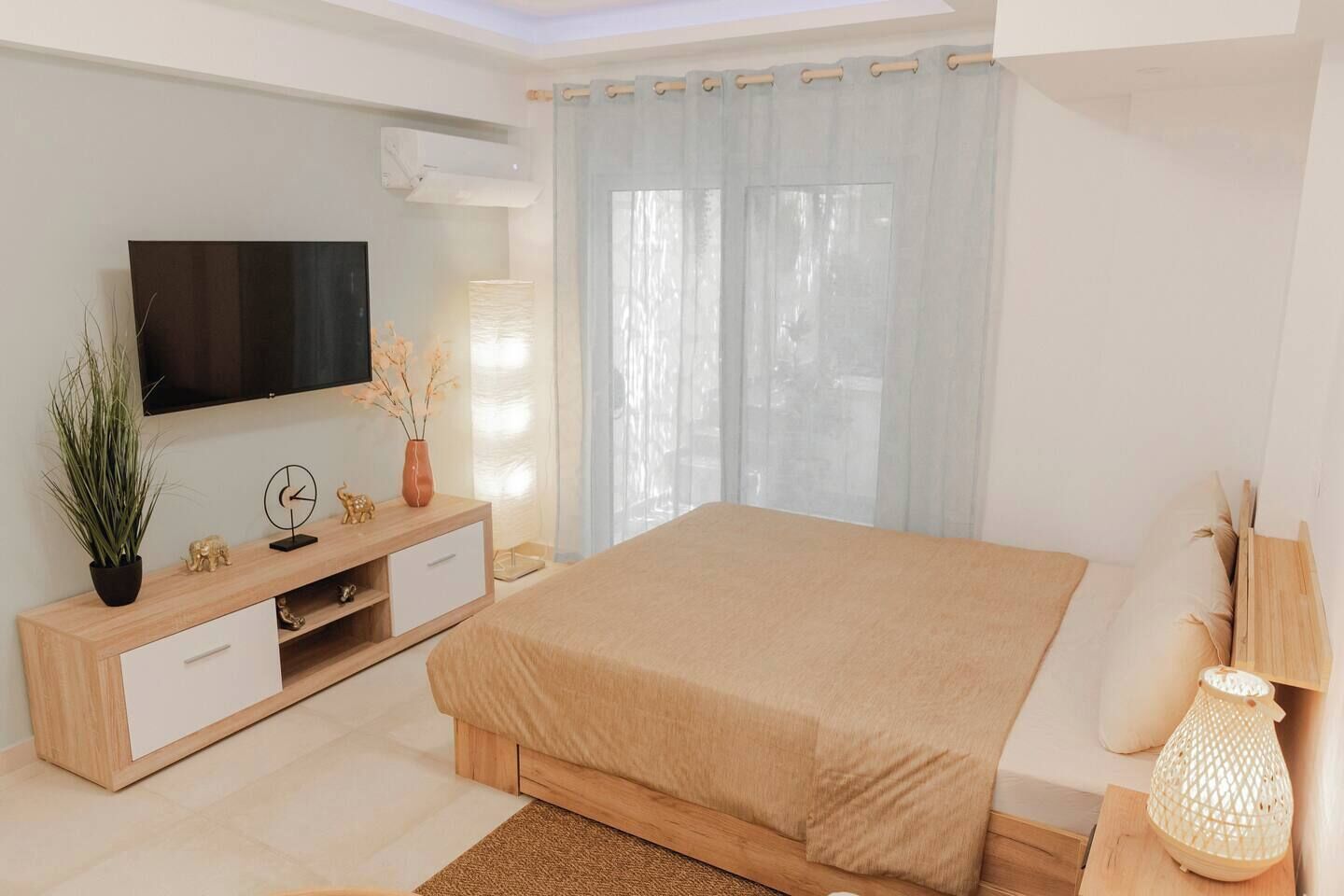 A bedroom with a bed, a TV, a wooden dresser, a plant, and a window with curtains.