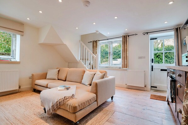 A modern living room with a beige sofa, a small ottoman, and a wooden floor. There is a staircase leading to an upper level, and large windows with curtains. - Cottage in Painswick Near Cotswold Way (Stroud)