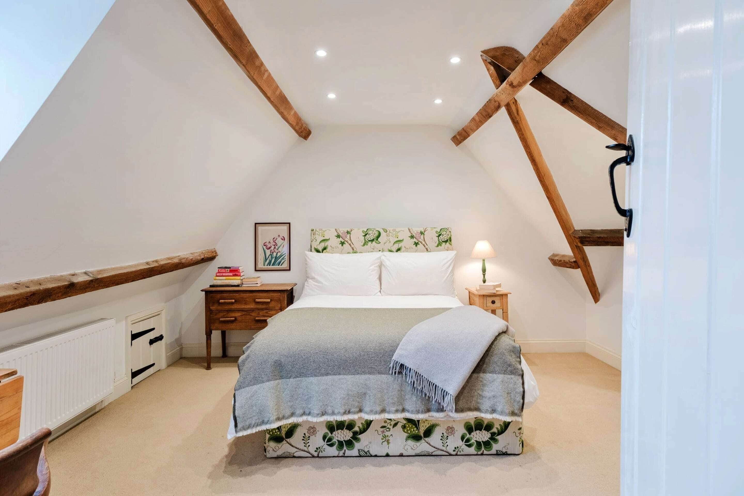 An attic bedroom with a sloped ceiling, wooden beams, a bed with a floral-patterned bedspread, a nightstand with a lamp, and a framed picture on the wall.