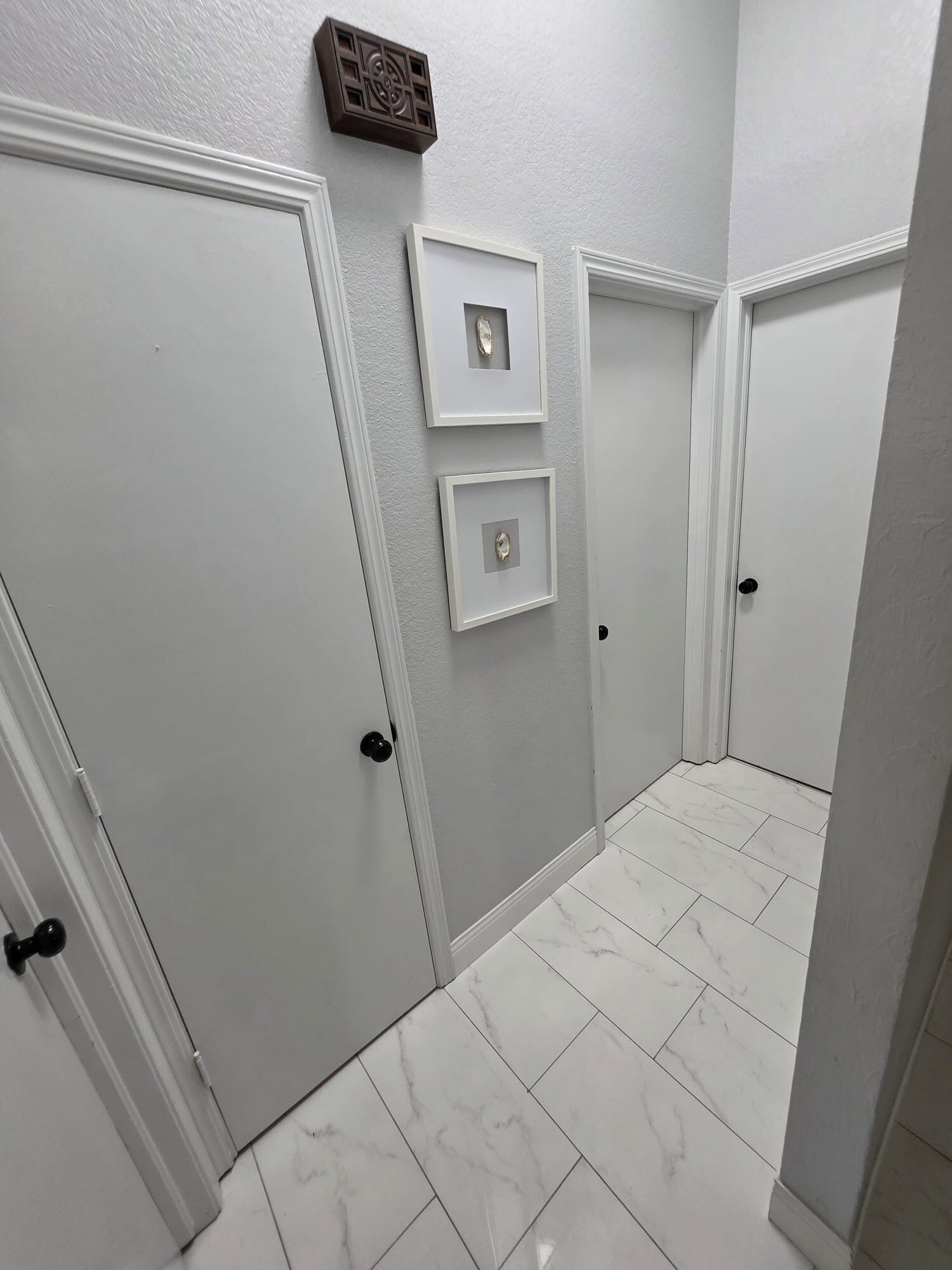 Un couloir avec des portes blanches, un sol en marbre et deux tableaux encadrés accrochés au mur.