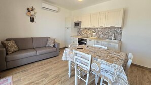 A compact living space with a sofa, a dining table with chairs, a kitchen area with cabinets and a microwave, and a decorative wall piece. - Home Geranio With Private Garden (Castelsardo)
