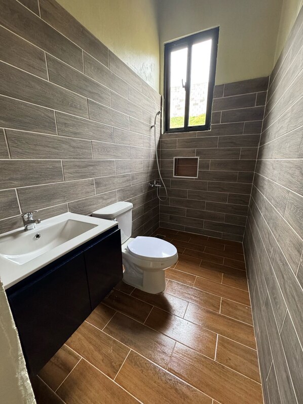 Une salle de bain avec des toilettes blanches, un lavabo blanc et une douche. Les murs sont recouverts de carreaux imitant le motif du bois, et le sol est en bois.