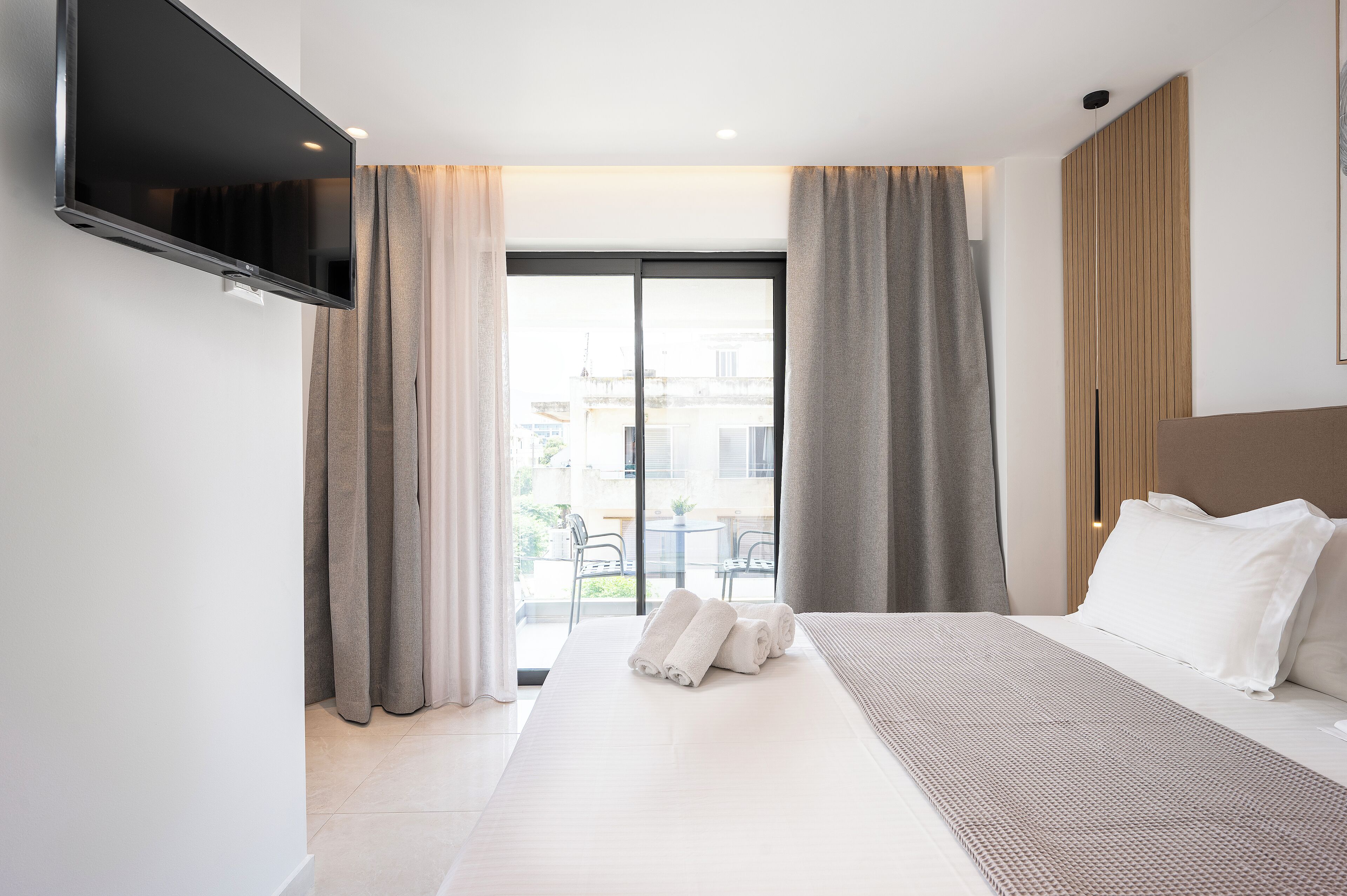 A modern hotel room with a large bed, a flat-screen TV mounted on the wall, and a balcony with a view of the city.