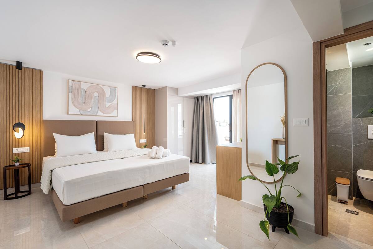 Una habitación de hotel moderna con una cama grande, una mesita de noche con lámpara, una planta en maceta y un baño con espejo e inodoro.
