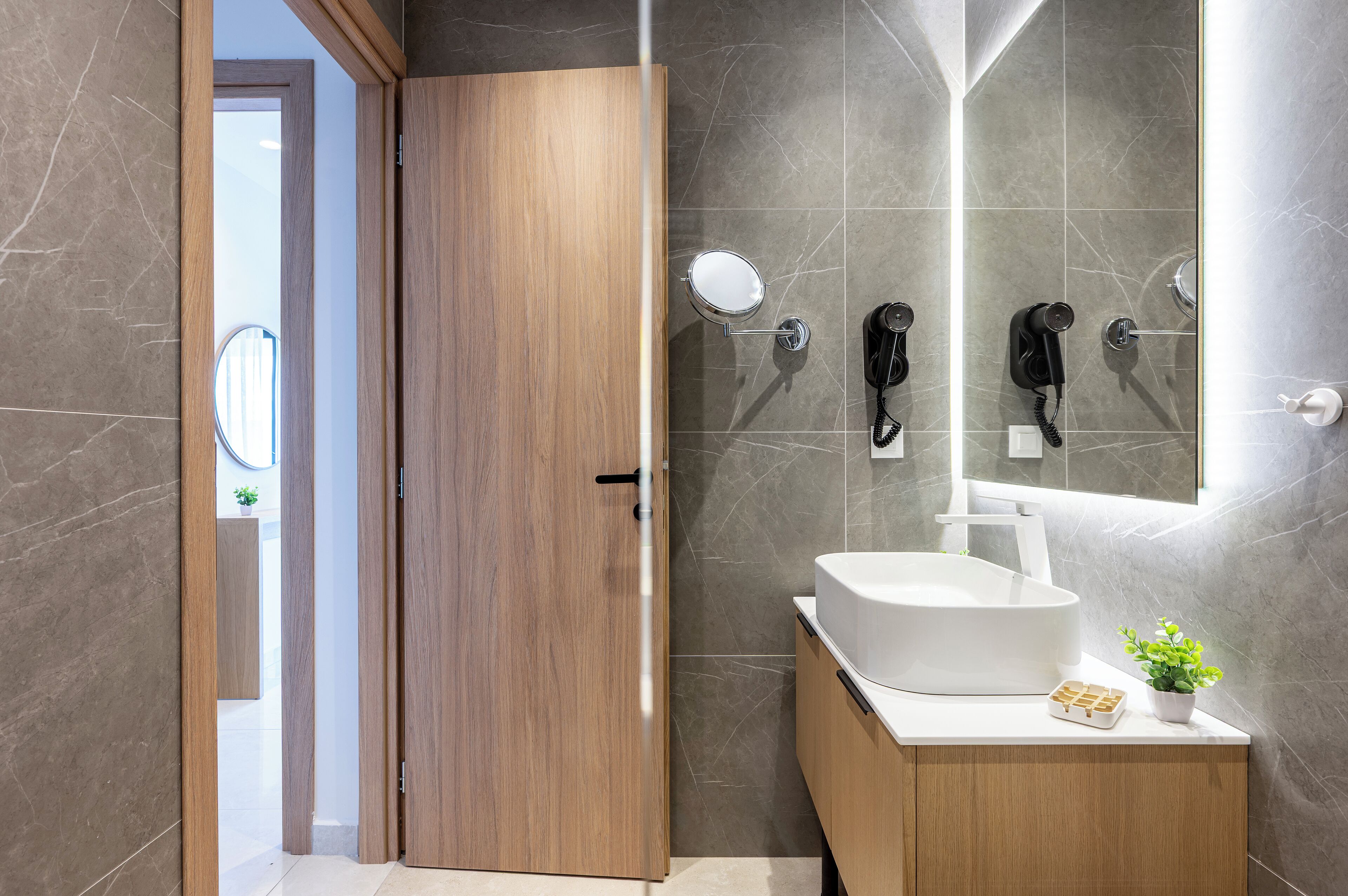 A modern bathroom with a wooden door, a white sink, and two hair dryers mounted on the wall.