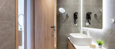 A modern bathroom with a wooden door, a white sink, and two hair dryers mounted on the wall.