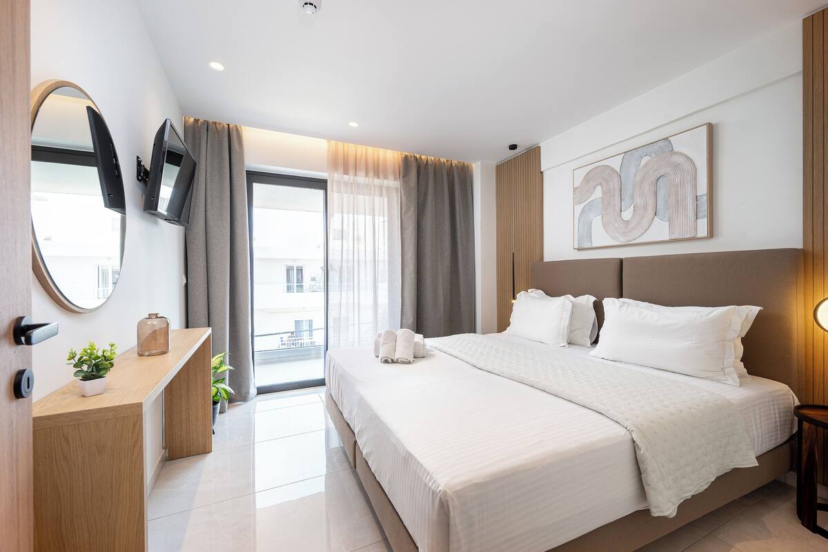 Habitación de hotel moderna con una cama grande, un escritorio con una planta, un televisor montado en la pared y un amplio ventanal con cortinas.
