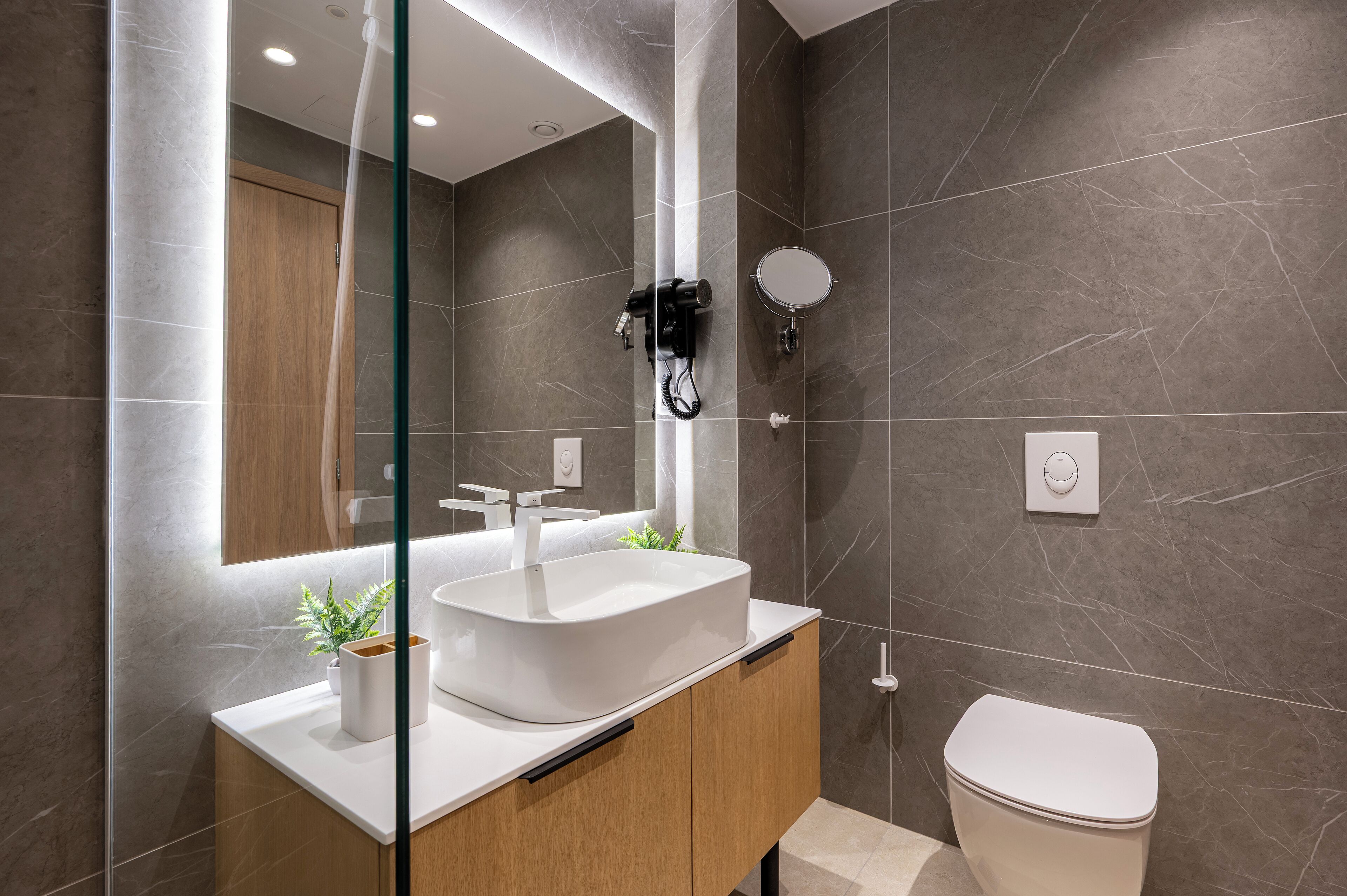 A modern bathroom with a large mirror, a white sink, a hairdryer, and a toilet.