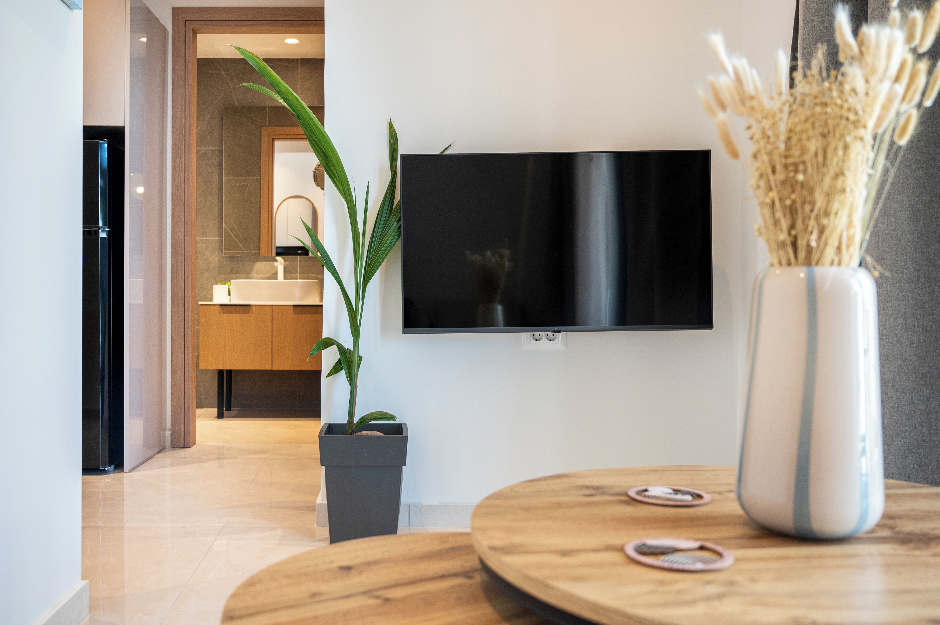 Ein modernes Wohnzimmer mit flachbildfernseher an der Wand, einem runden Couchtisch aus Holz, einem Blumentopf und einer Vase mit Trockenblumen.