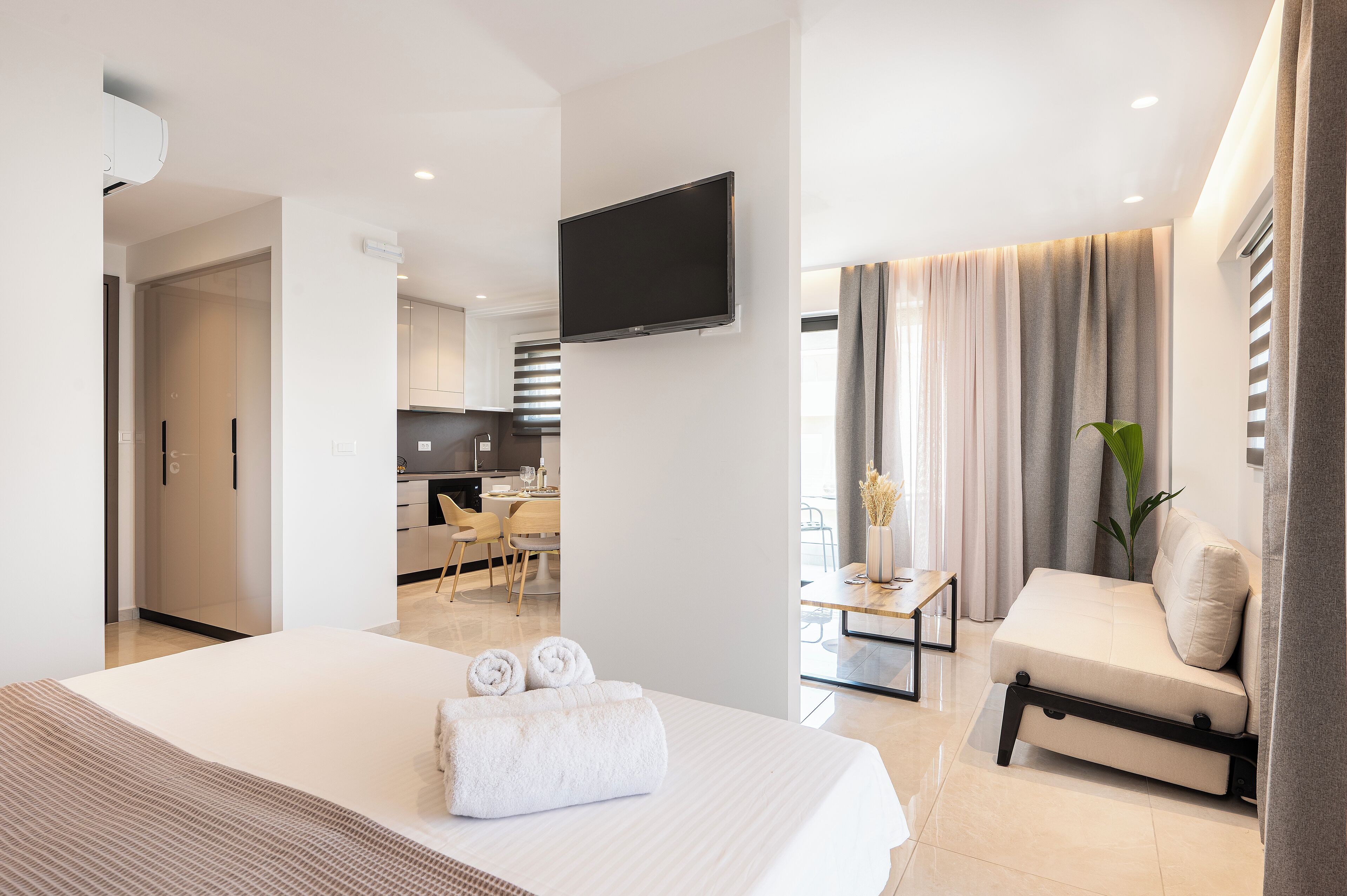 A modern hotel room with a large bed, a flat-screen TV, a sofa, and a dining area with a table and chairs.