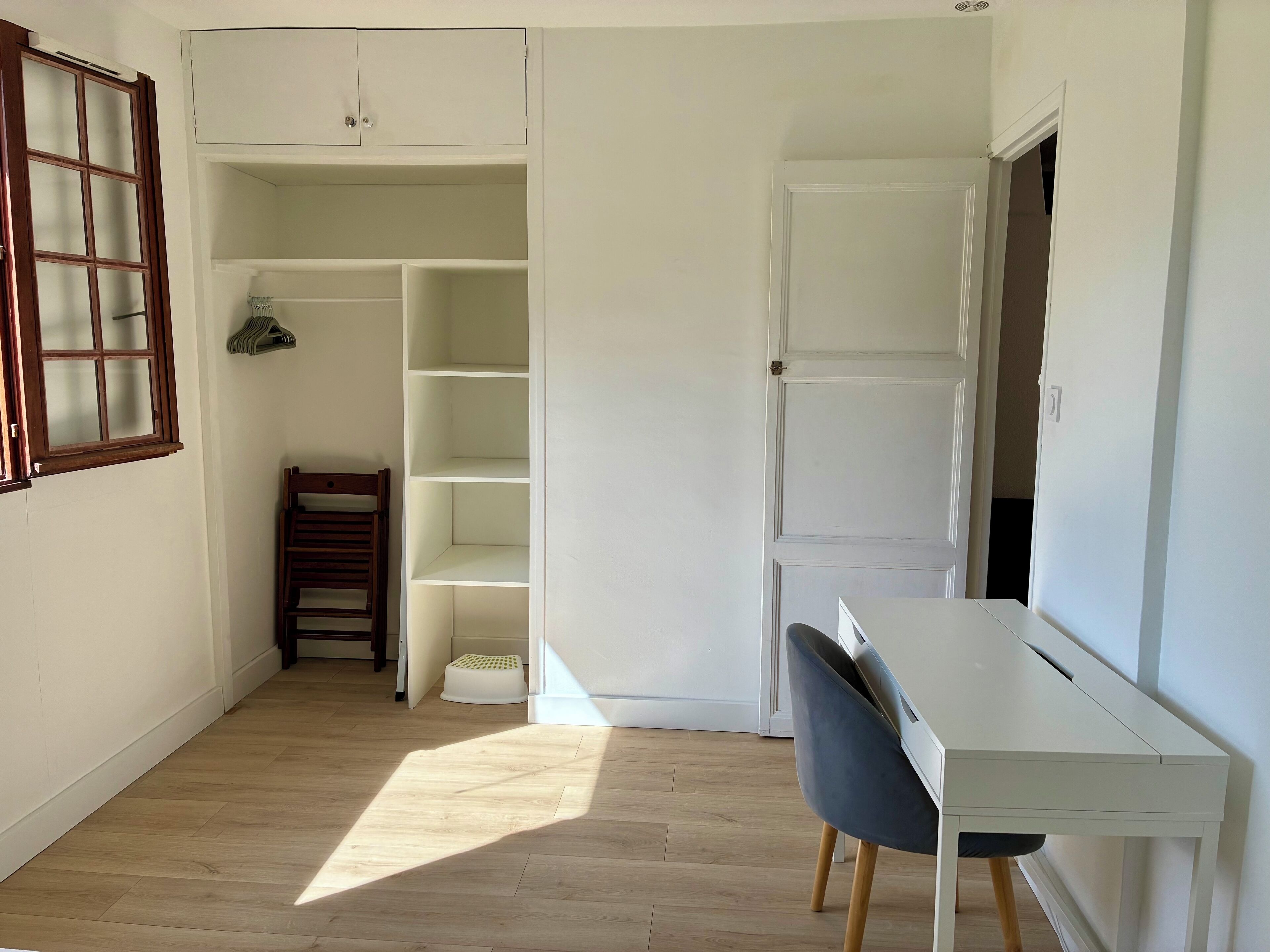 A room with a white desk, a chair, a wooden chair, a wardrobe, and a window.