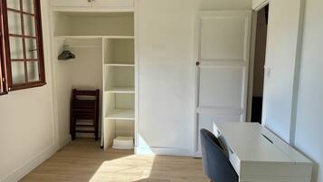 A room with a white desk, a chair, a wooden chair, a wardrobe, and a window.