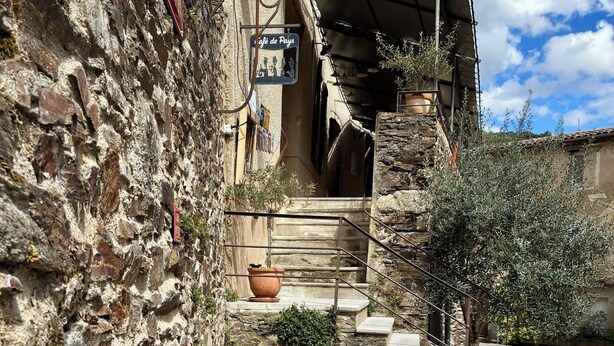 Ein rustikales Steinhaus mit einem kleinen Wasserspiel im Freien, Stufen, die zu einem überdachten Eingang führen, und einem Schild, das ein Café anzeigt.