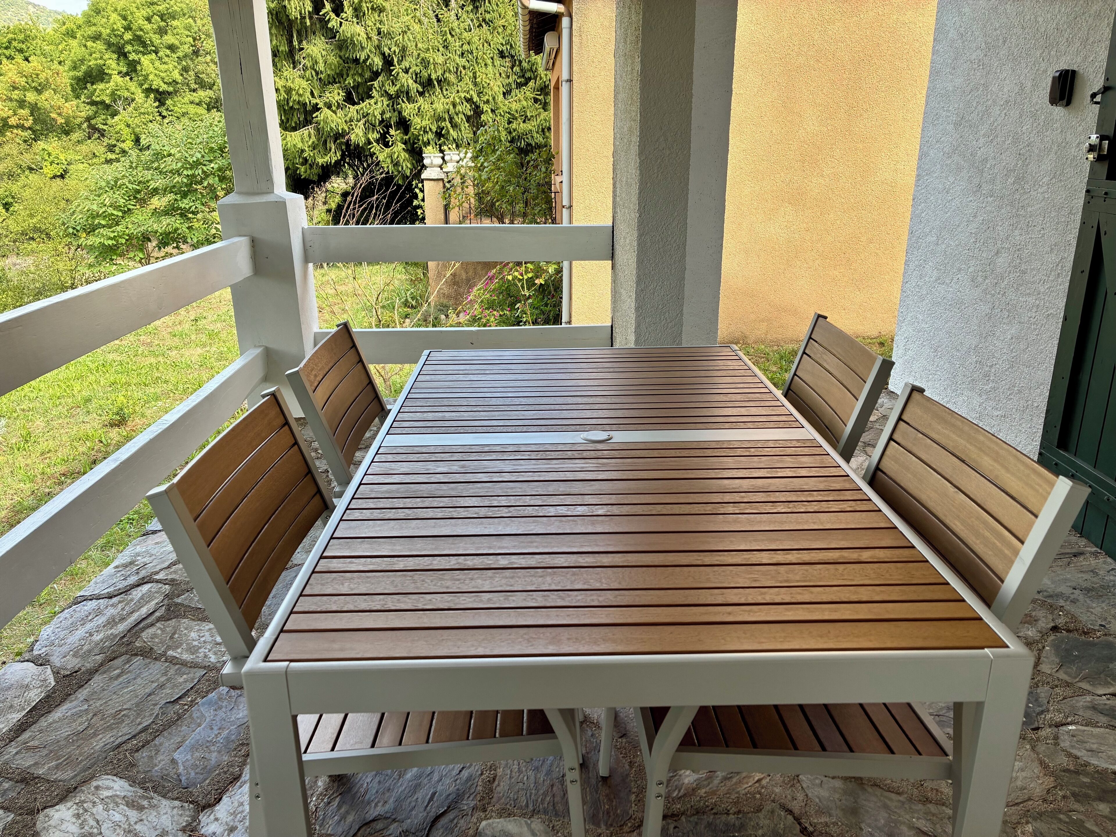 Una mesa de madera con cuatro sillas, ubicada en un patio empedrado con barandilla blanca.