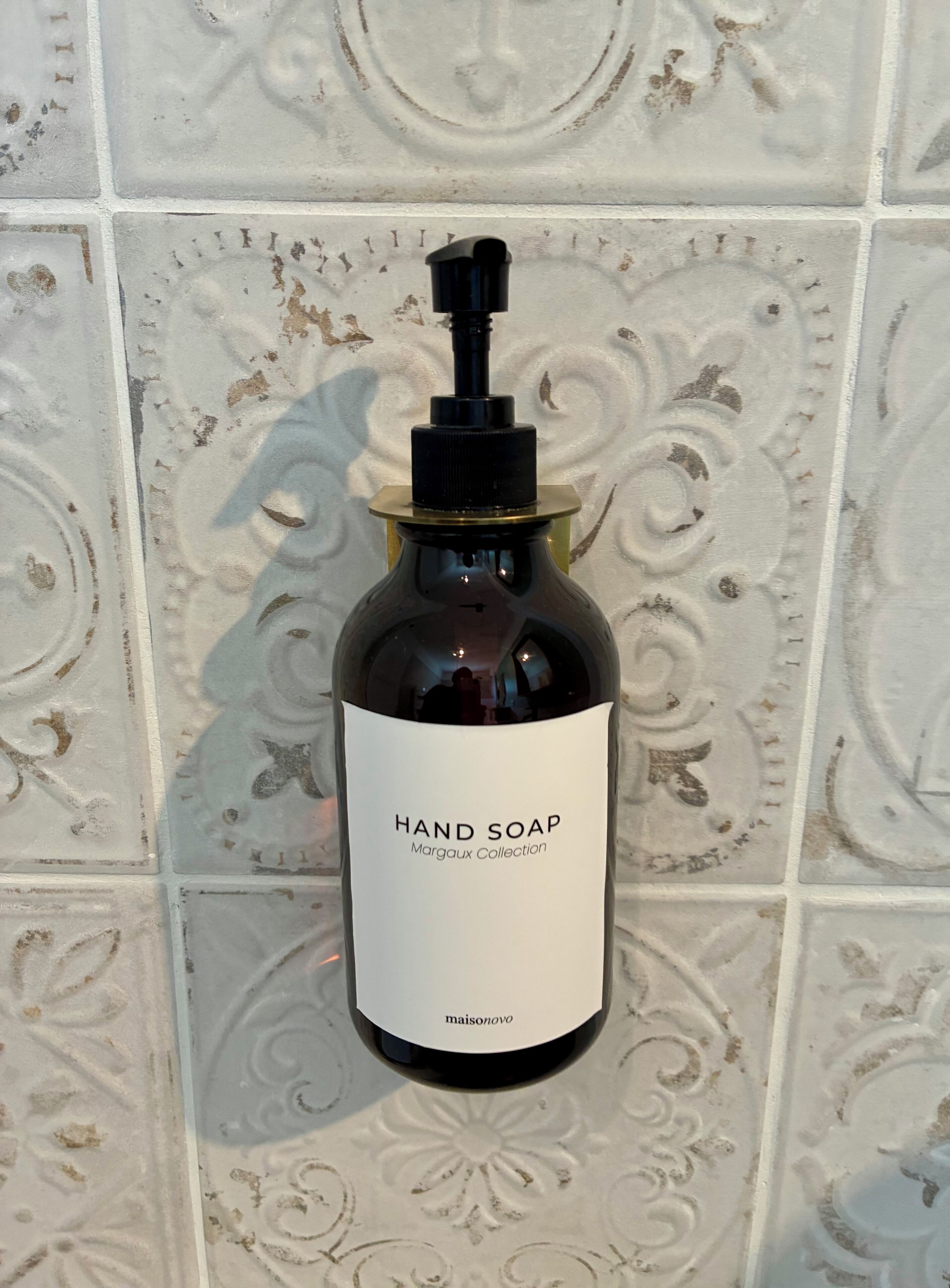 Una ampolla de sabó de mans amb l'etiqueta 'Hand Soap Margaux Collection' penjada en una paret de rajoles.
