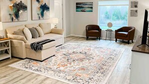 Ein Wohnzimmer mit einem beige gefärbten Sofa, zwei Sesseln, einem Couchtisch, einem großen Teppich, Deckenventilatoren und Kunstwerken an den Wänden.