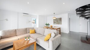 A modern living room with a beige sectional sofa, a wooden coffee table, and a dining area with a table and chairs. There is a black spiral staircase and a painting on the wall.